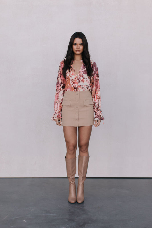 Woman wearing a floral blouse, beige skirt, and tan boots against a plain background