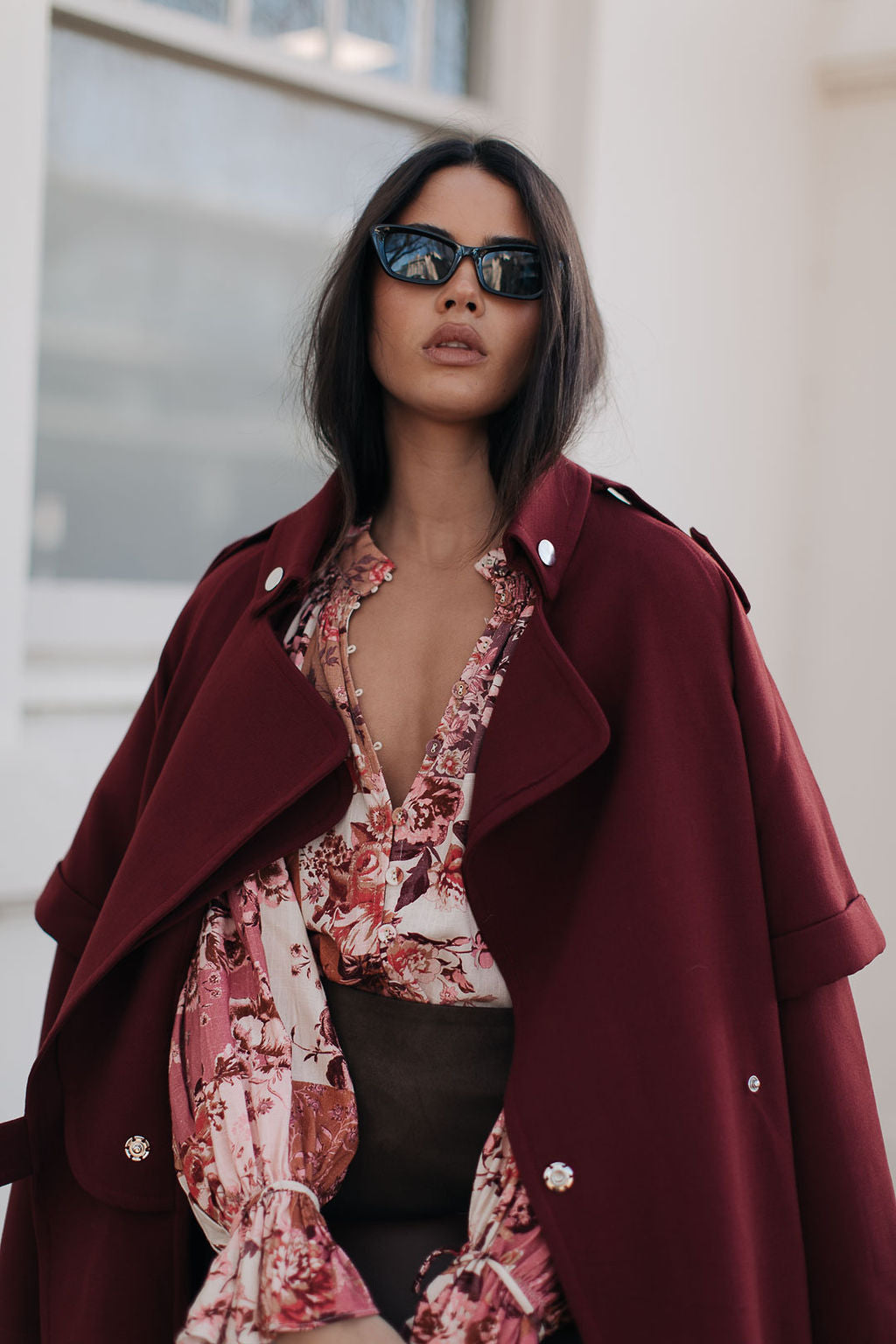 Woman wearing a burgundy coat over a floral dress with sunglasses, standing outdoors.
