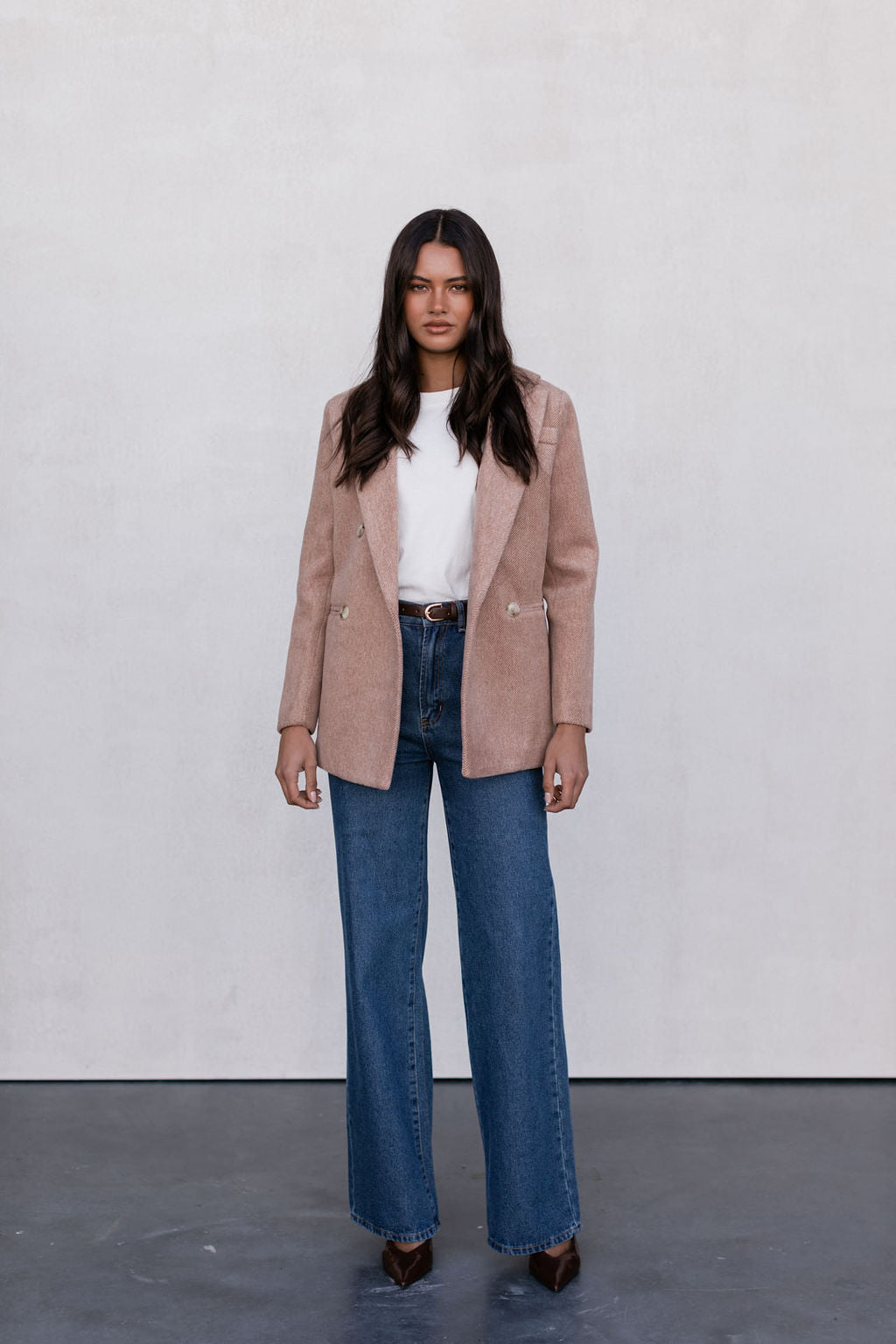 Woman wearing a beige blazer, white top, and blue jeans against a plain background