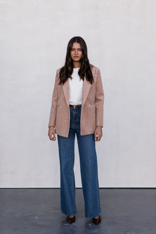 Woman wearing a beige blazer, white top, and blue jeans against a plain background