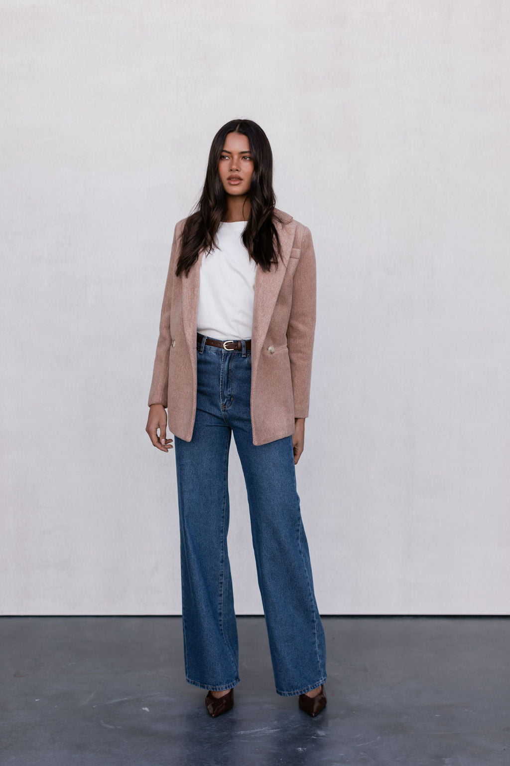 Woman wearing a beige blazer, white shirt, and blue jeans against a plain background