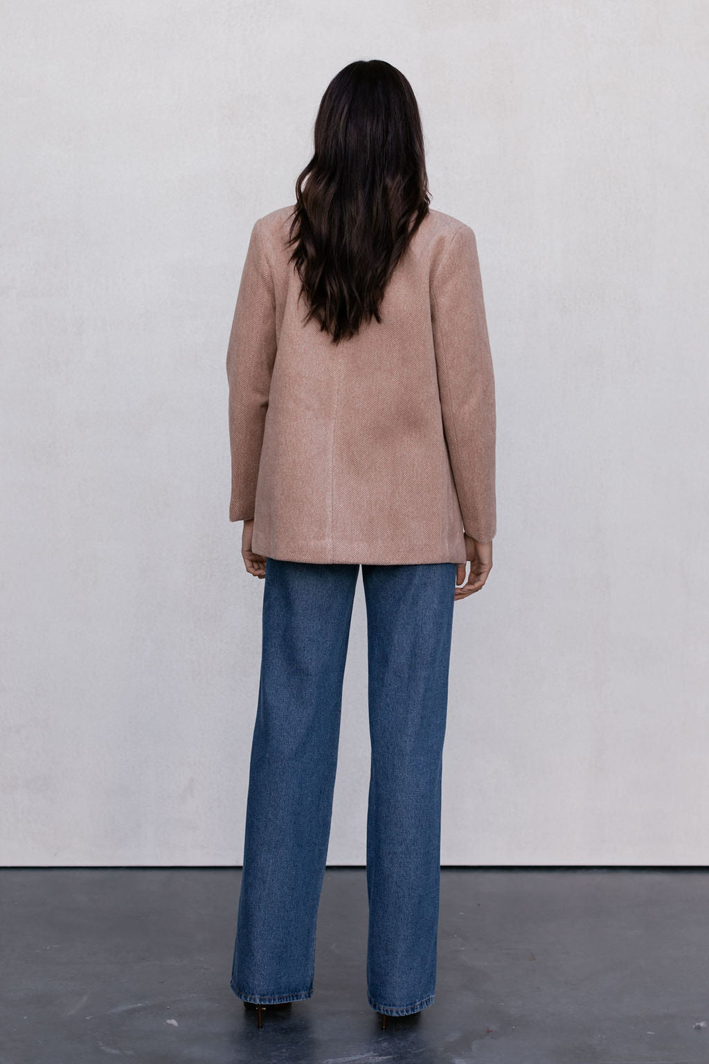 Person wearing a beige coat and blue jeans standing against a white wall.