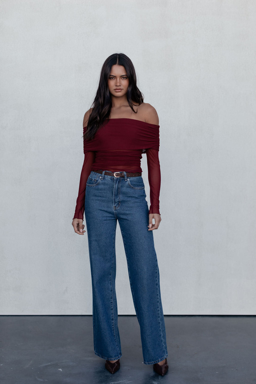 Woman wearing a burgundy off-shoulder top and blue jeans against a plain background