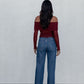 Woman wearing a red off-shoulder top and blue jeans against a white wall.