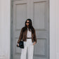 Woman in a brown jacket and white pants standing in front of a gray door.