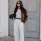 Woman in a stylish outfit with a brown jacket, white shirt, and white pants standing against a gray door.