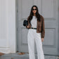 Woman in a stylish outfit with a brown jacket, white pants, and black clutch standing in front of a gray door.