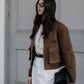 Woman wearing a brown jacket and white pants against a gray wall.