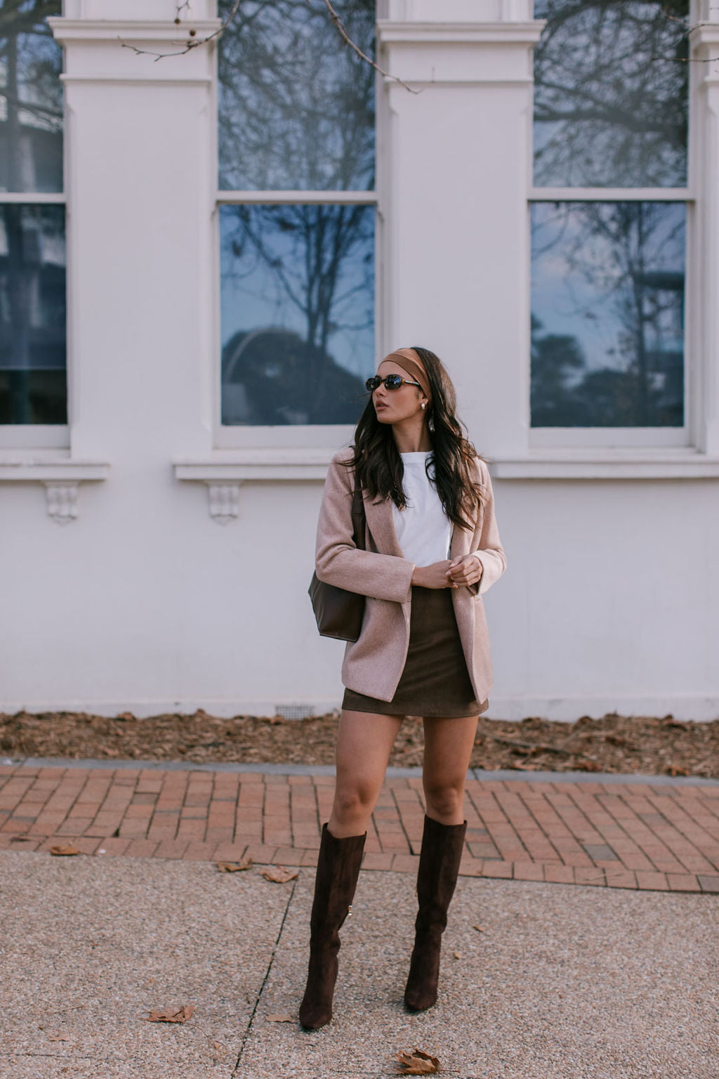 Woman in a beige coat and brown knee-high boots standing in front of a white building.