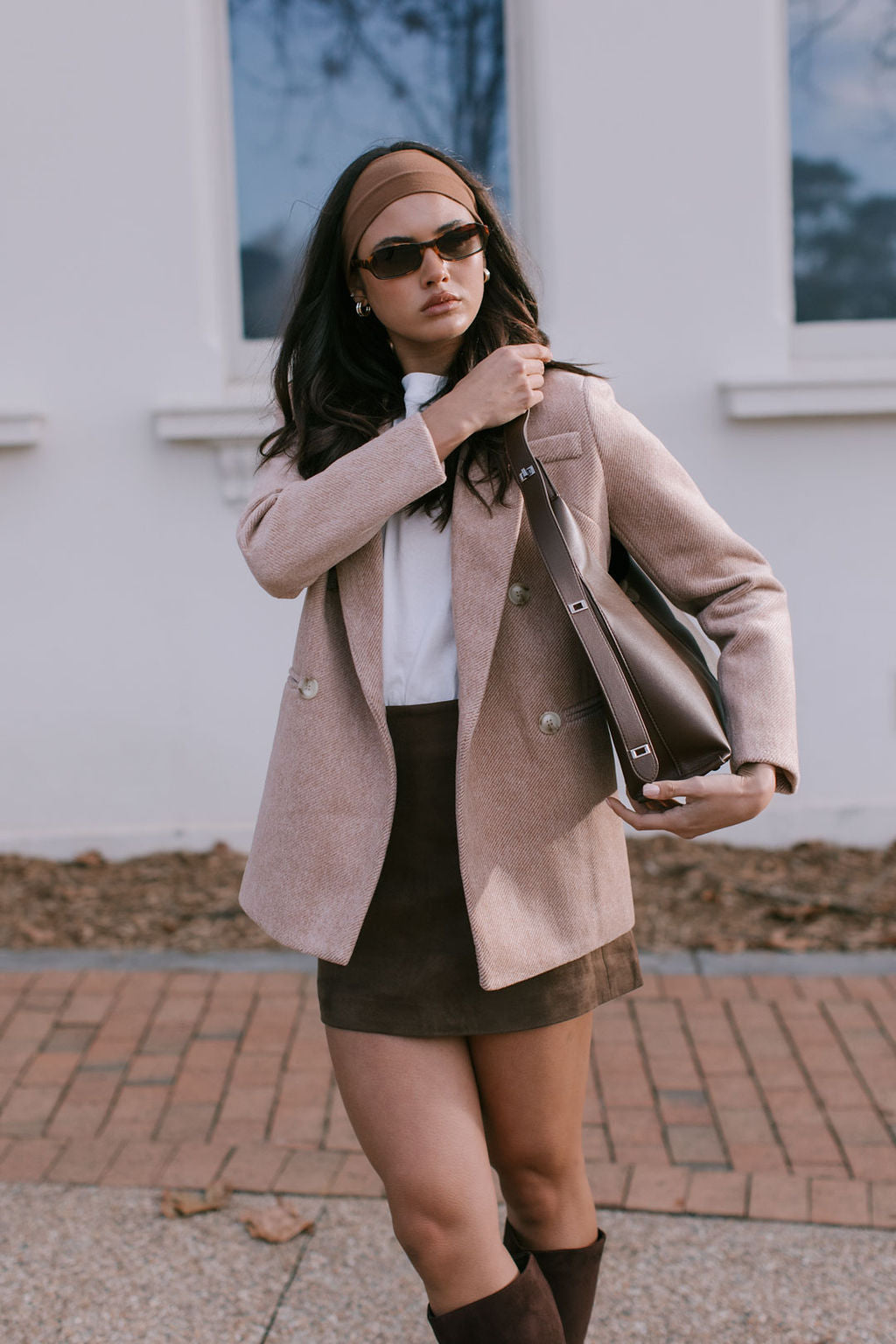 Woman in a beige coat and brown skirt holding a handbag outdoors.