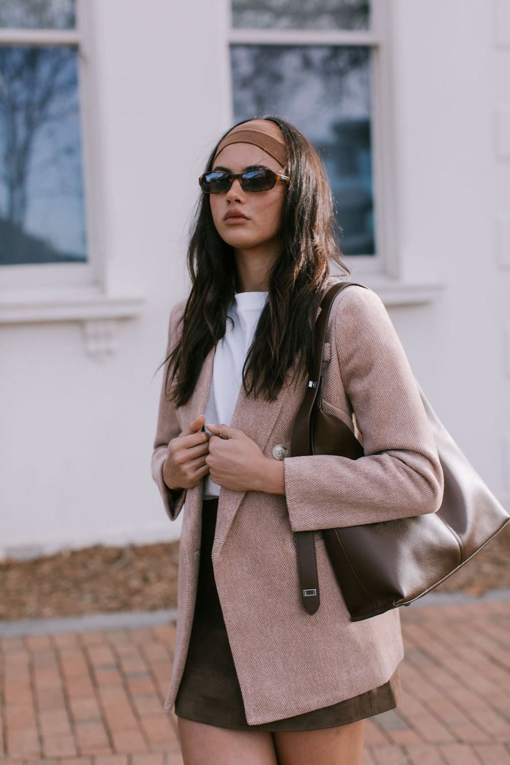 Woman styled wearing a beige blazer coat, brown mini skirt, headband and sunglasses outdoors.