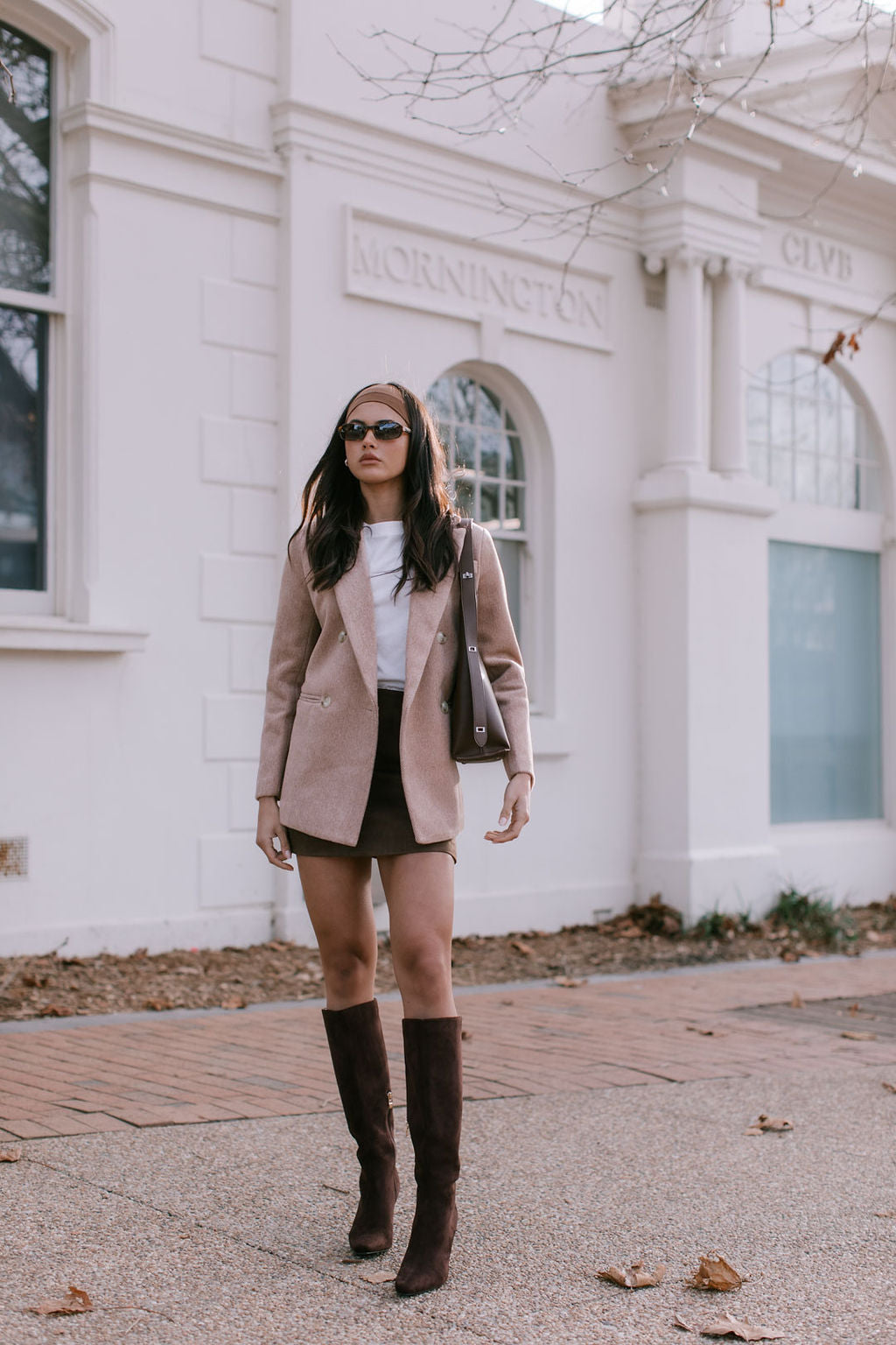 Woman in a beige coat and brown knee-high boots standing in front of a white building.