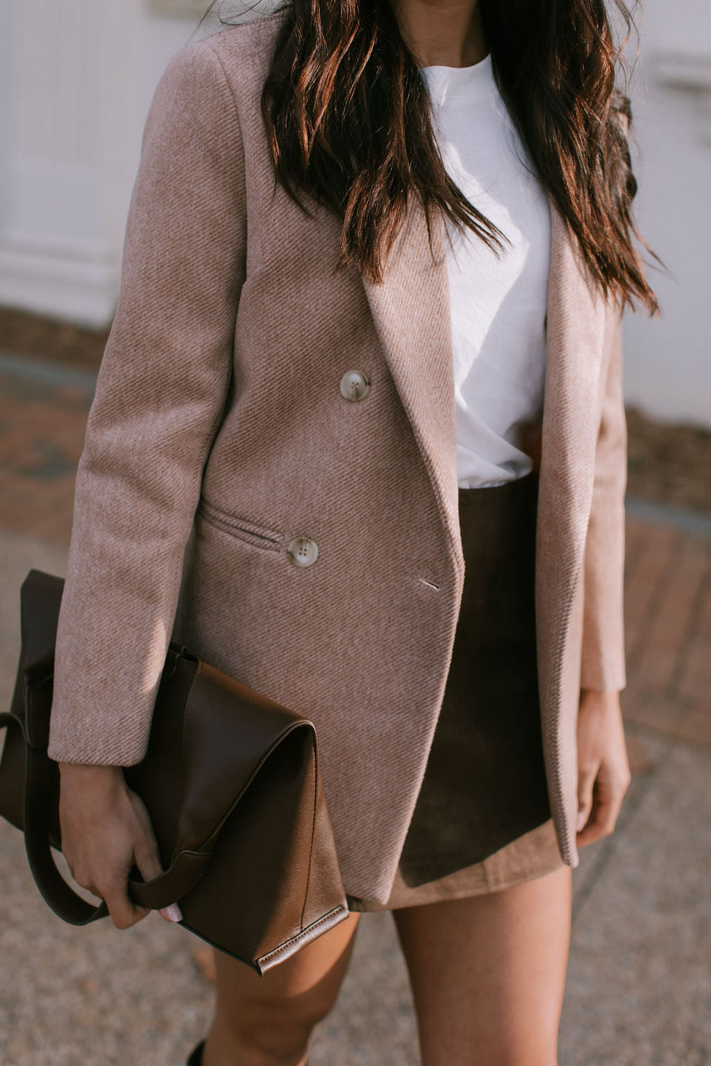Person wearing a brown beige blazer coat and holding a brown bag on a blurred background