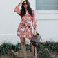 Woman in a floral dress and knee-high boots holding a brown leather bag against a white wall.