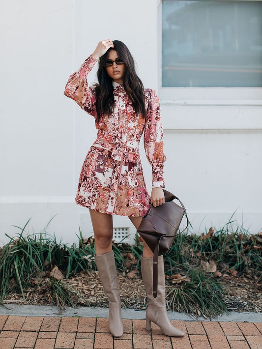 Woman in a floral dress and knee-high boots holding a brown leather bag against a white wall.