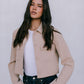 Woman wearing a beige jacket over a white shirt and dark jeans against a plain background