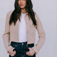 Woman wearing a beige jacket over a white shirt and dark jeans against a plain background