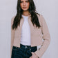 Woman wearing a beige jacket over a white shirt and dark jeans on a plain background