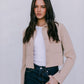 Woman wearing a beige jacket, white shirt, and dark jeans against a plain background