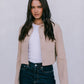Woman wearing a beige jacket, white shirt, and dark jeans against a plain background
