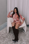 Woman in a floral dress and knee-high boots sitting on a white chair.