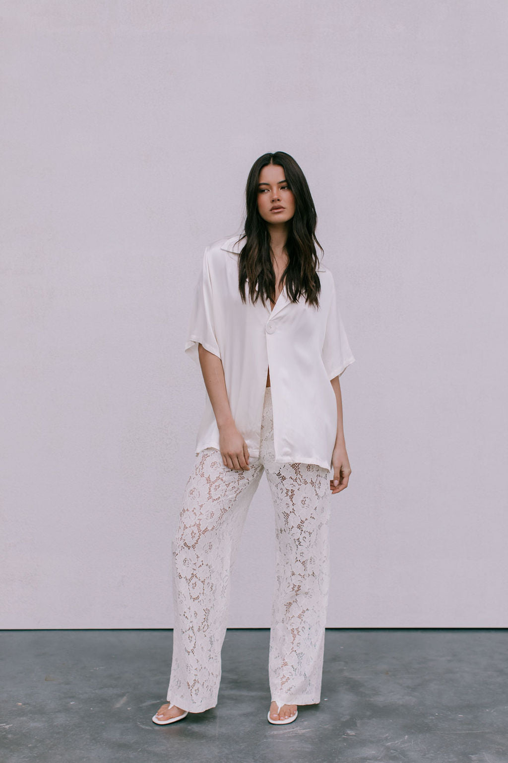 Woman wearing a white shirt and lace pants against a plain background