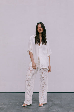 Woman wearing a white shirt and lace pants against a plain background