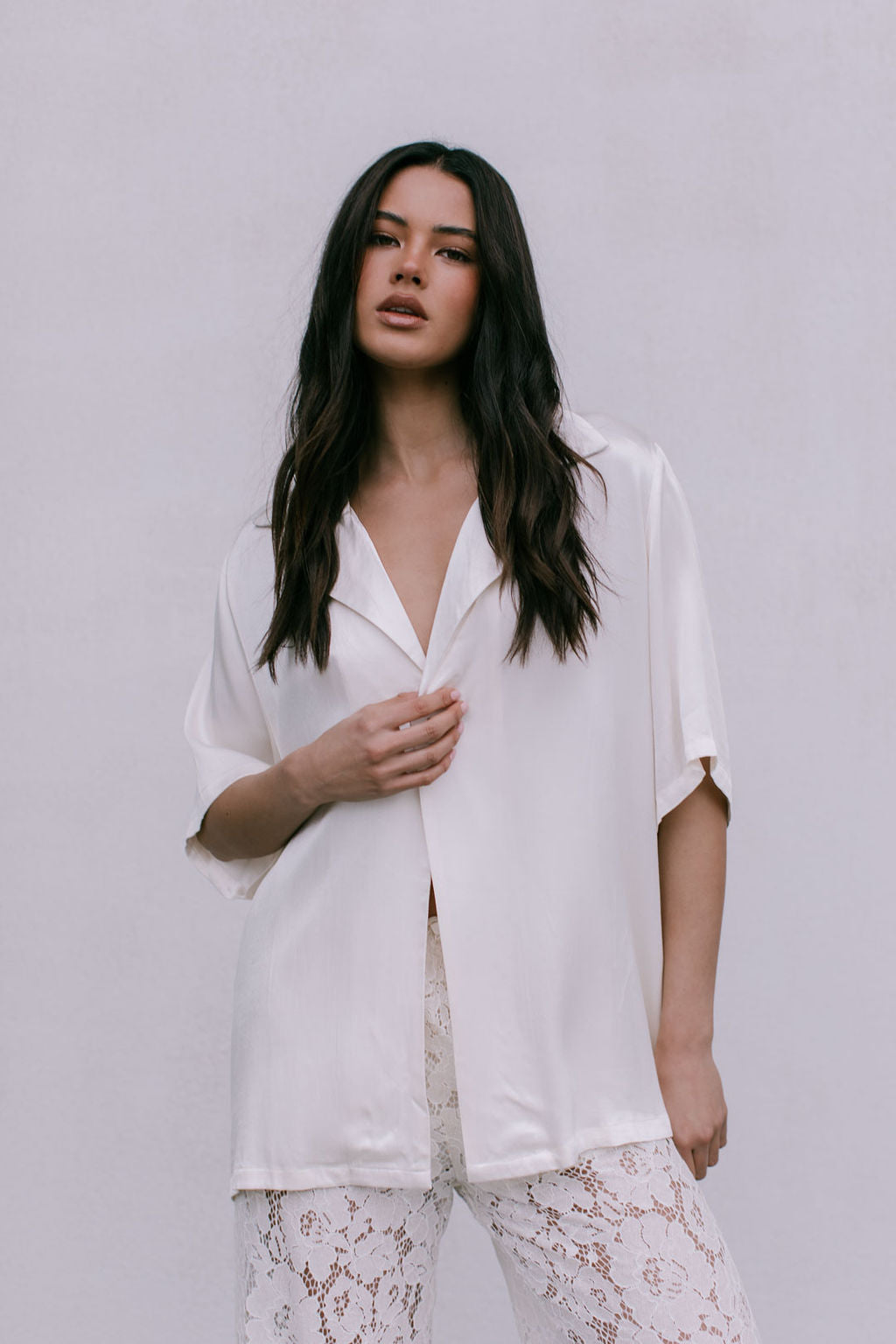 Woman wearing a white blouse with a deep neckline and lace pants on a plain background