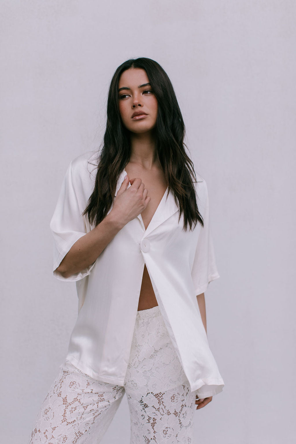 Woman wearing a white satin shirt and lace pants against a plain background