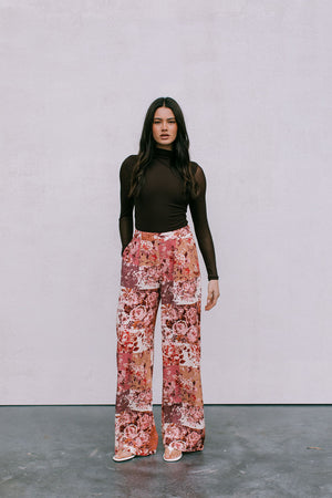 Woman wearing a brown top and floral pants against a plain background