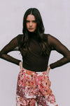 Woman wearing a brown mesh top and floral pants against a plain background