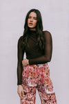 Woman wearing a brown long-sleeve top and floral pants against a plain background