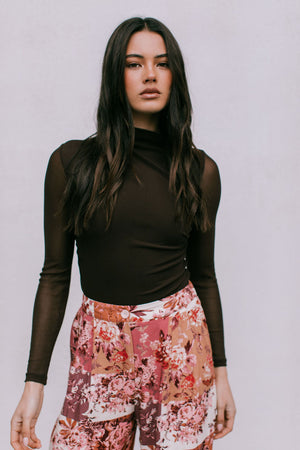 Woman wearing a brown long-sleeve top and floral pants against a plain background