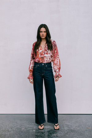 Woman wearing a floral blouse and dark jeans against a plain background