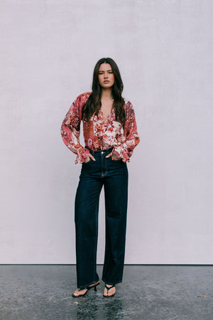 Woman wearing a floral blouse and dark jeans against a plain background