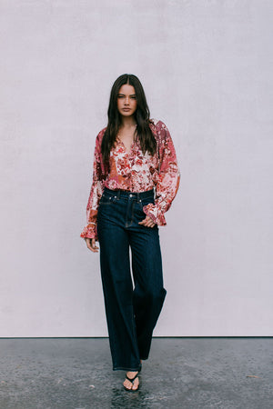 Woman wearing a floral blouse and dark jeans against a plain background