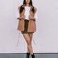 Woman wearing a beige vest over a white shirt and brown skirt with black boots on a plain background
