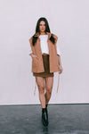 Woman wearing a tan quilted vest, white top, brown skirt, and black boots on a plain background