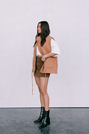 Woman wearing a tan vest, white shirt, brown skirt, and black boots against a white background