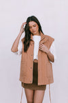 Woman wearing a tan vest over a white shirt and brown skirt on a white background