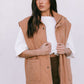 Woman wearing a tan vest over a white shirt and brown skirt on a plain background