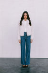 Woman wearing a white fur vest and blue jeans against a plain background