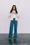 Woman wearing a white fur coat, white shirt, and blue jeans against a plain background