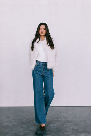 Woman wearing a white fur coat, white shirt, and blue jeans against a plain background