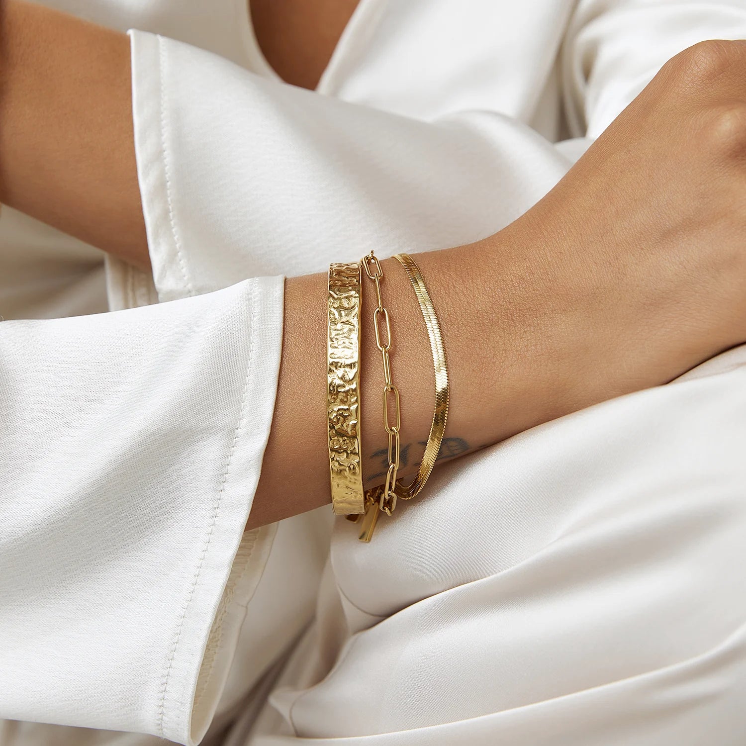 A close-up of a woman wearing gold bracelets, styled as a stack with a mix of chains and cuffs.